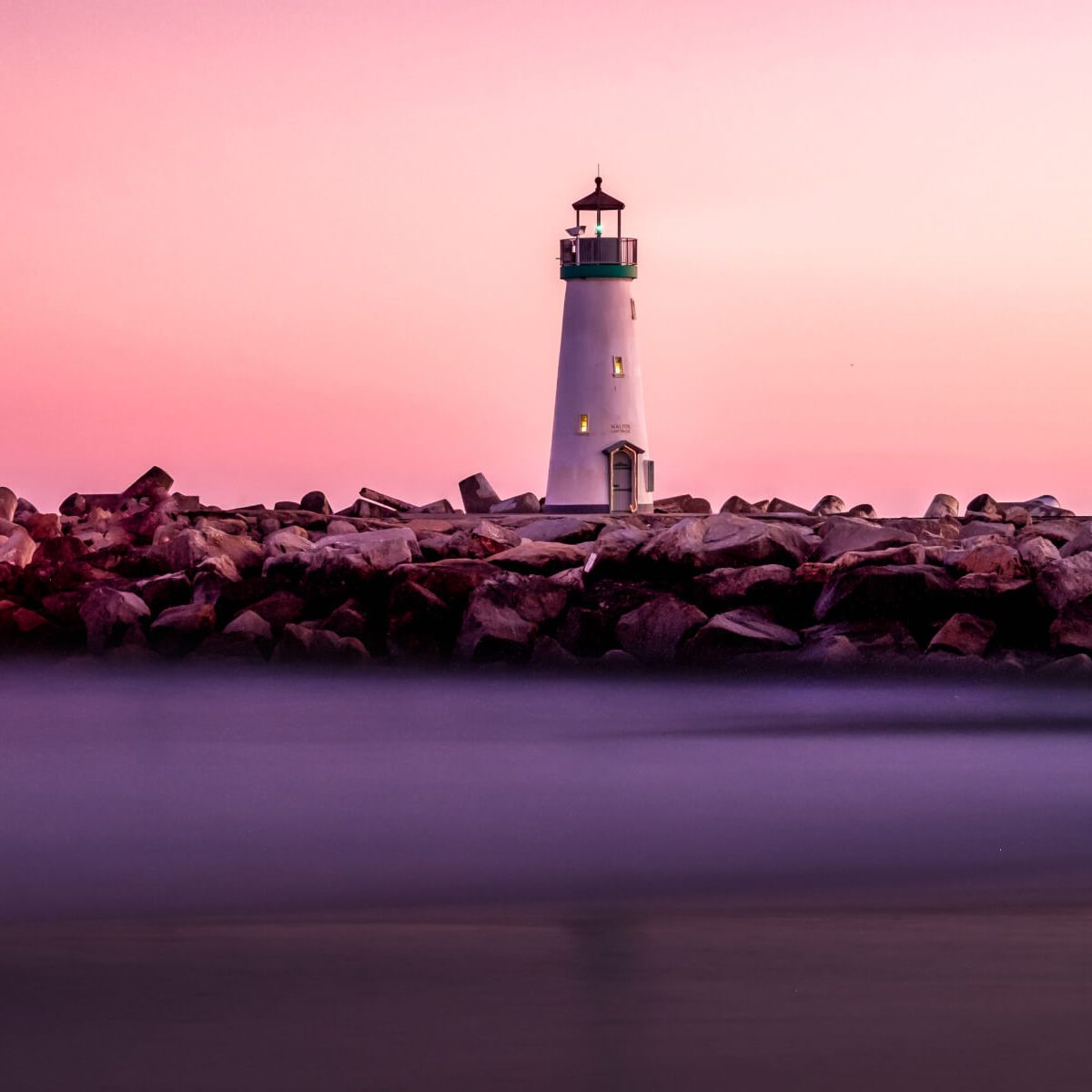 A lighthouse on a rocky shore, with purple water reflecting a rose sunset sky.
