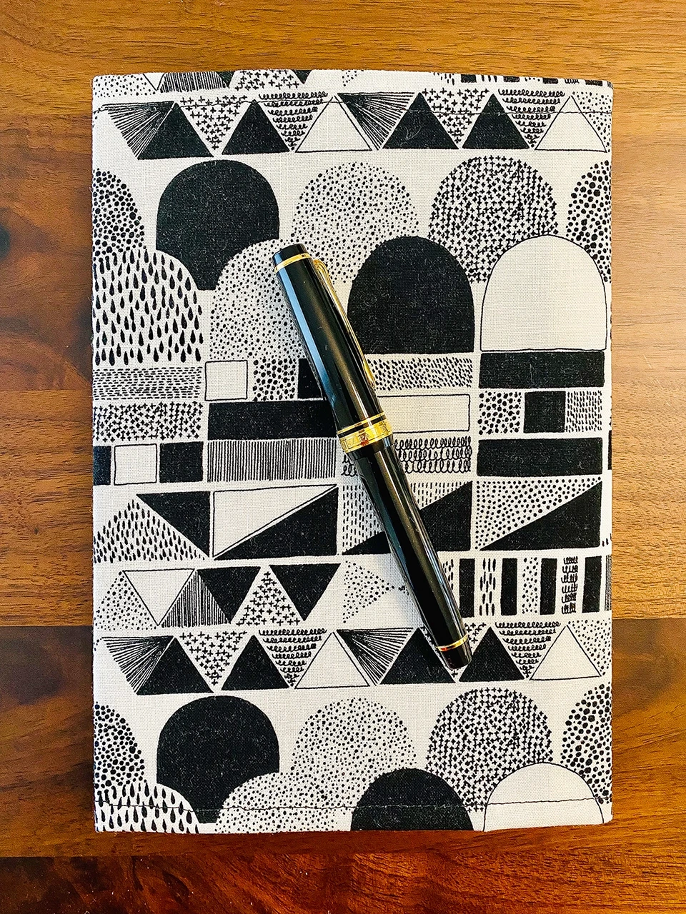 An A5 journal clad in a single panel of fabric in abstract black and white doodled shapes. A black and gold Sailor fountain pen sits beside the journal