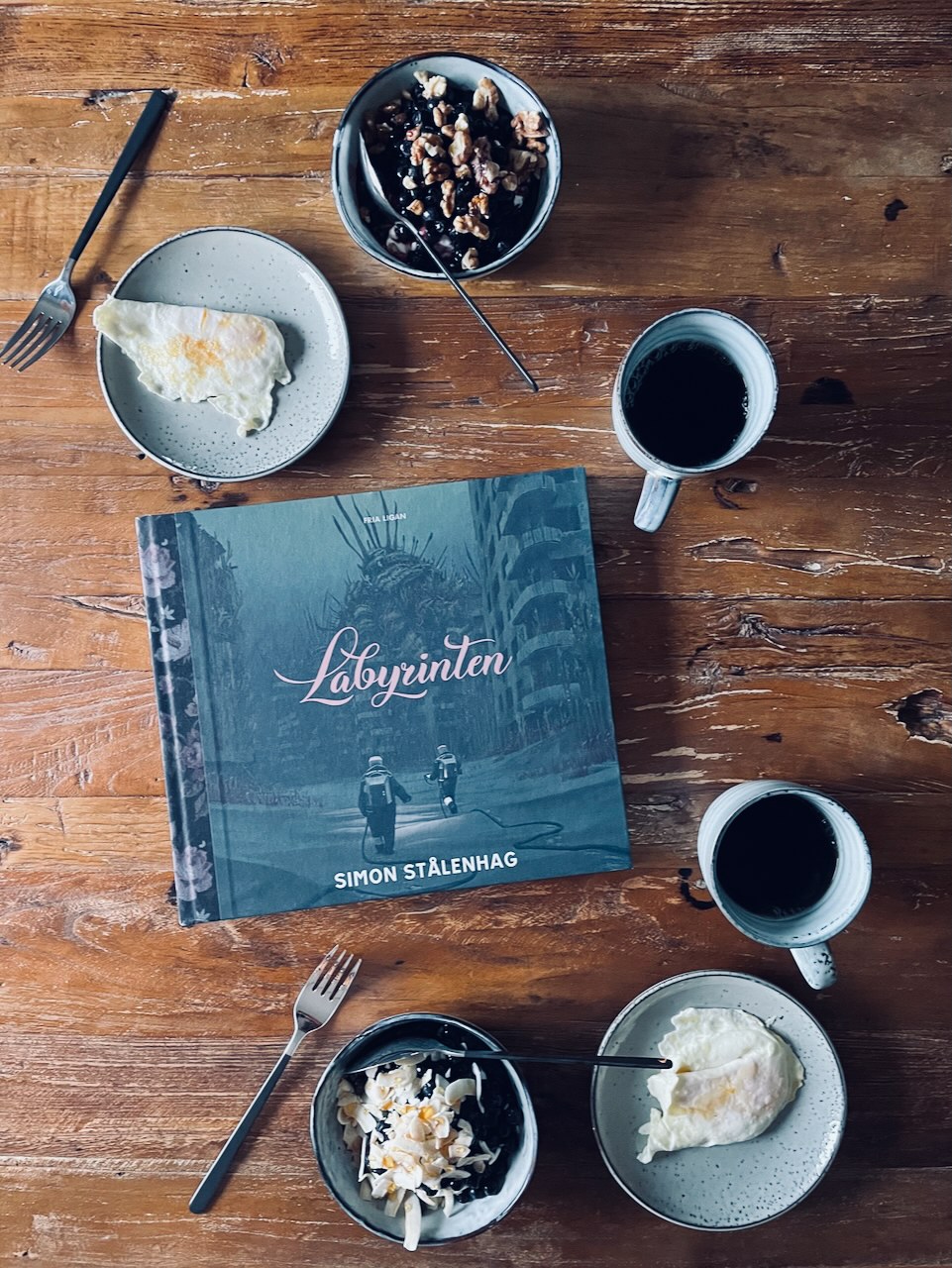 A breakfast table with eggs, coffee, and yogurt topped with blueberries. In the middle of the table is a book by Simon Stålenhag: The Labyrinth.