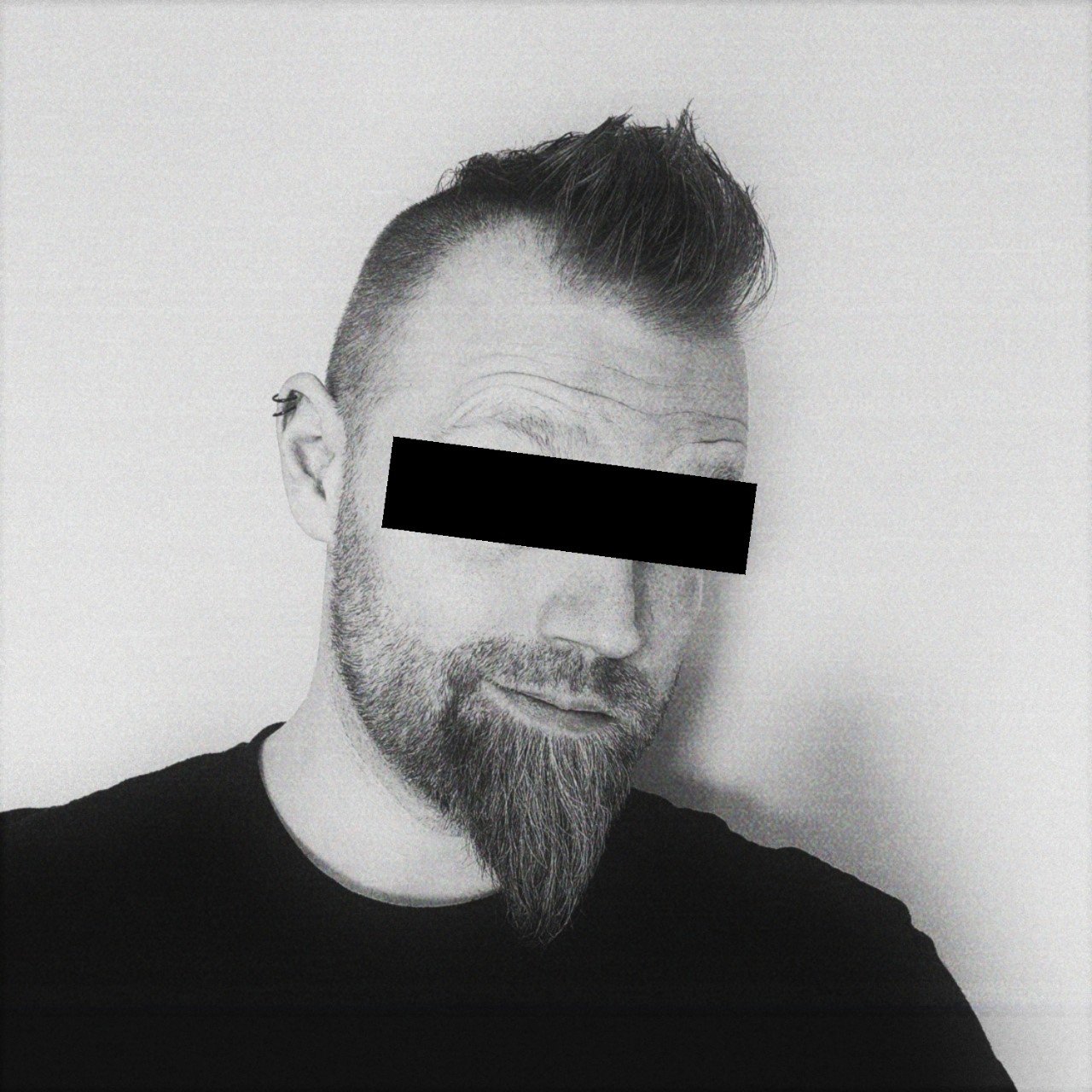 A black and white grainy photo of Myles's head and shoulders. He has a long pointy beard and a mohawk, with a black censored bar across his eyes