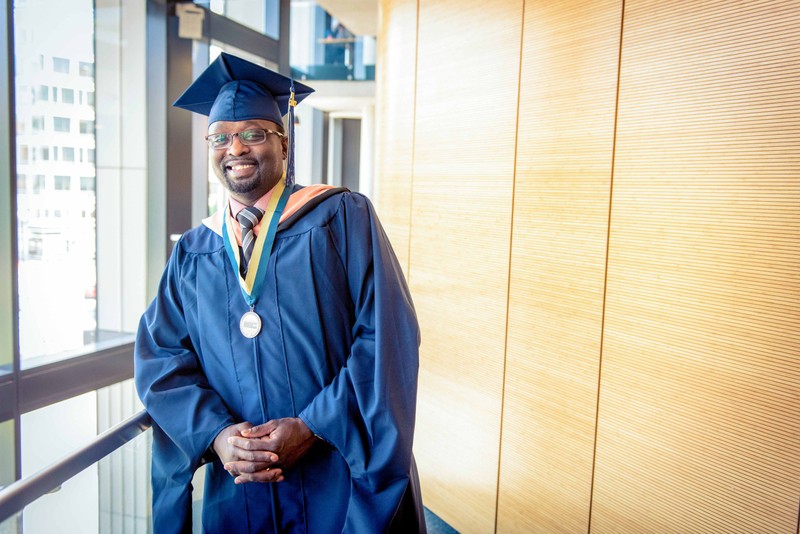 A smiling George Washington University graduate in his cap and gown with his hands folded