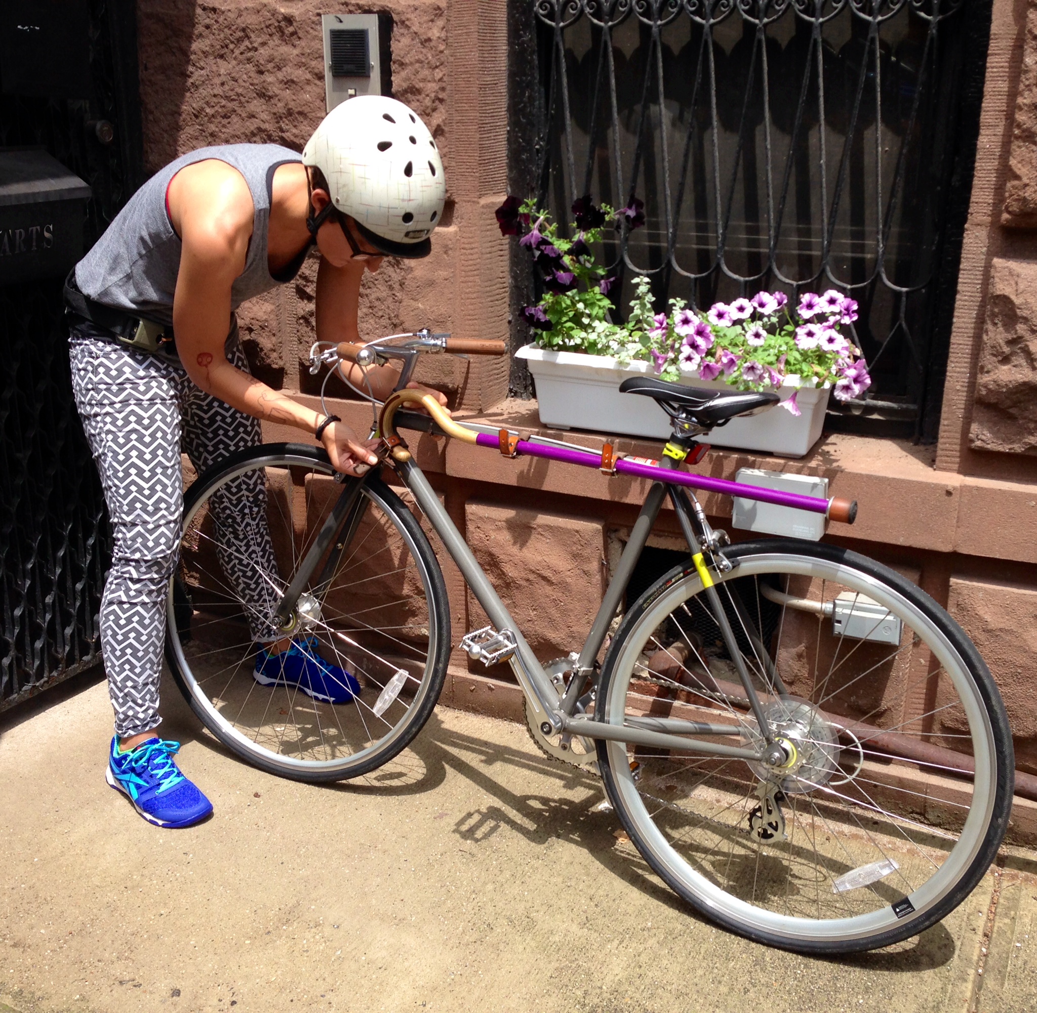Liz Jackson on a New York City sidewalk, locking up her bike. Her purple cane is attached by leather straps and metal buckles in parallel to the bike's long body, keeping it secure and out of the way for her ride.