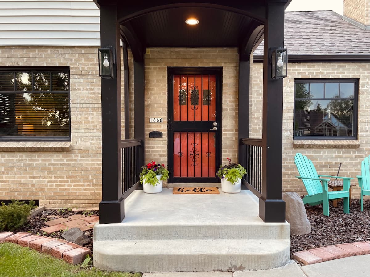 Brightly colored potted plants sit on either side of a reddish-orange front door and a welcome mat that says “Hello”