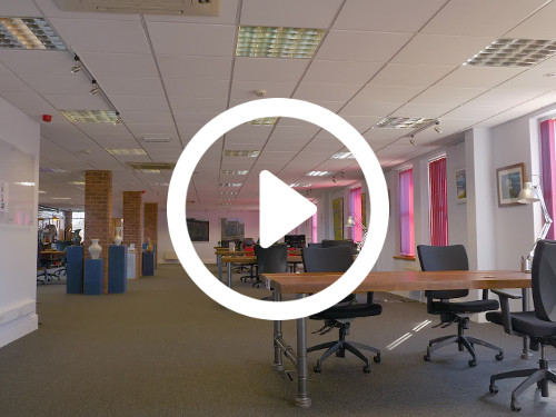 Spacious shared office workspace with ceiling tiles containing multiple wooden desks and black office chairs Skype background