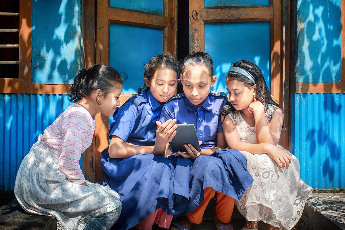Girls reading on a tablet outside of a school in Bangladesh as part of Concern Worldwide's CRAAIN (Collective Responsibility, Action and Accountability for Improved Nutrition) program