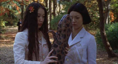 A screenshot from the comedy movie 'Pavillion Salamandre' of two women awkwardly holding a large salamander.