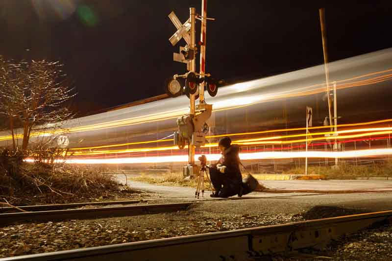 Molly kneeling in front a camera on a tripod next to railroad tracks with a long exposure light trails from a truck driving by.