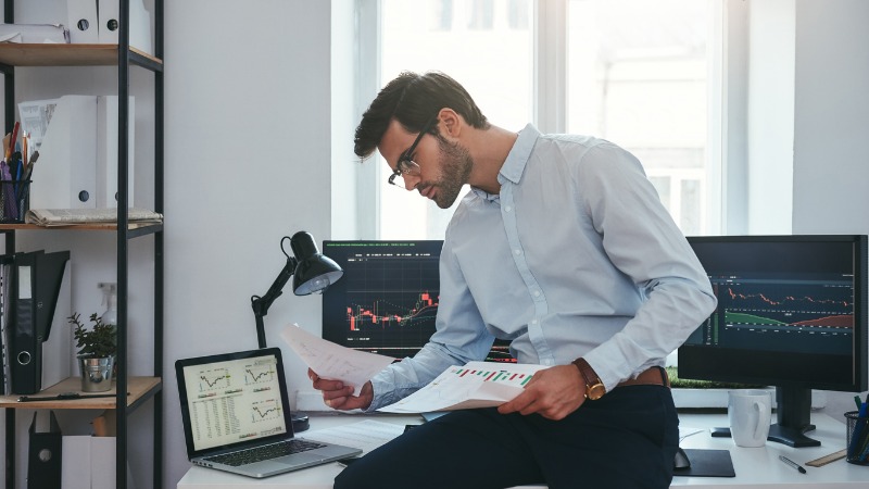 A worker studying printed analyses next to several computer screens with diagrams and reports.