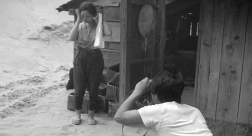 A black and white animated gif of a scene from the movie 'Woman in the Dunes' showing a man photographing a nervous woman in front of her house, surrounded by sand.