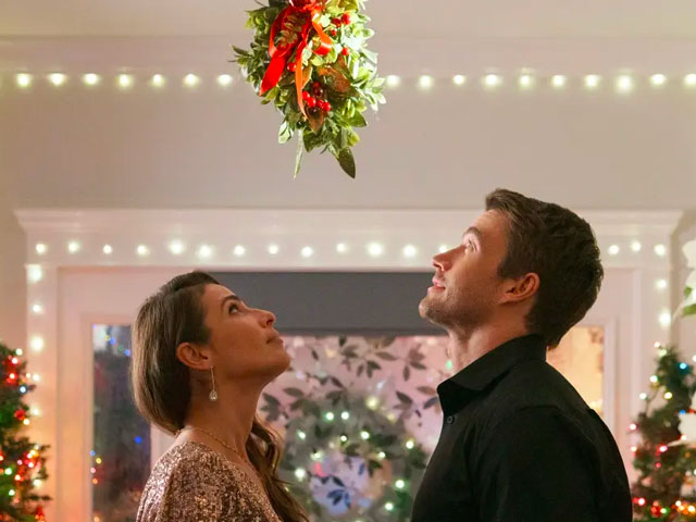 A man and a woman standing under the mistletoe, which means they have to drink in the Hallmark Christmas Movie Drinking Game