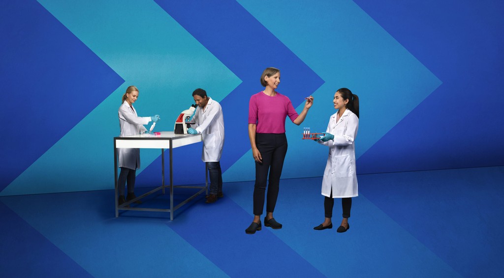 Three people in white lab coats and a woman in the pink shirt working in a stylized lab with chevron backdrops.