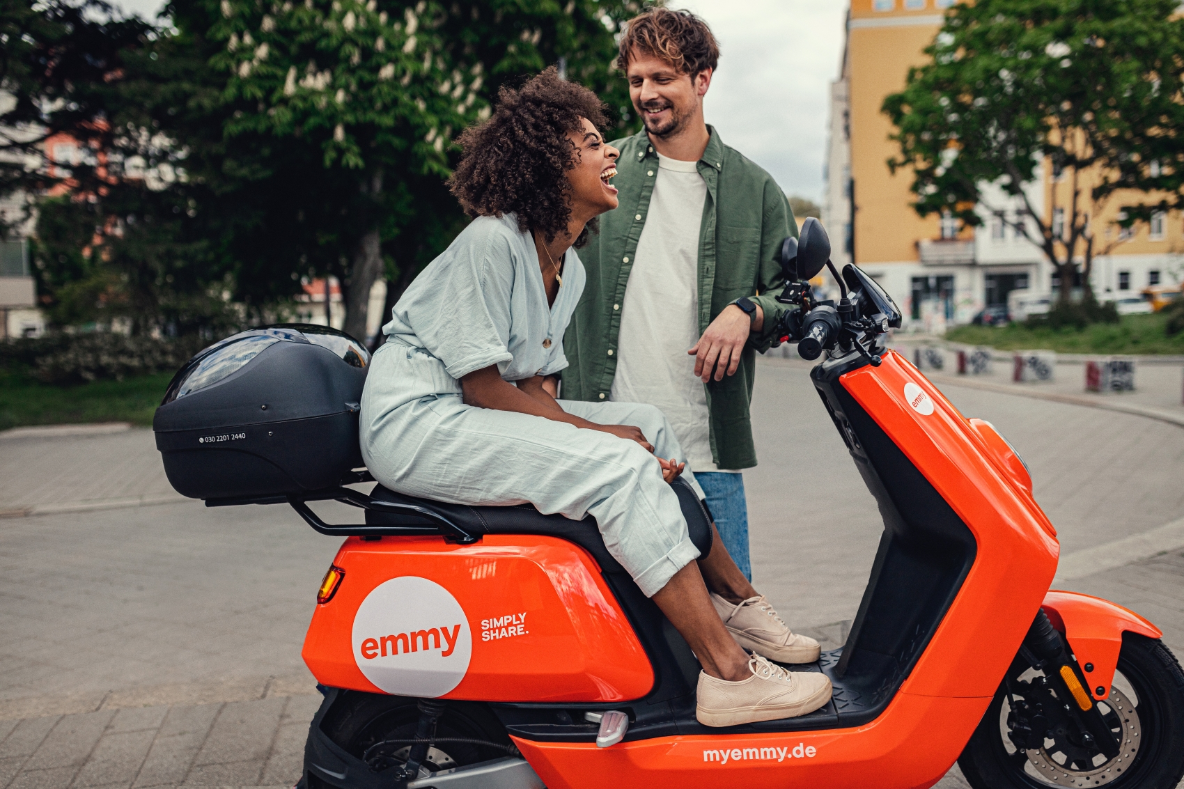 emmy customer card of a caucasian male standing next to an African American female person sitting on an emmy moped in the street. 