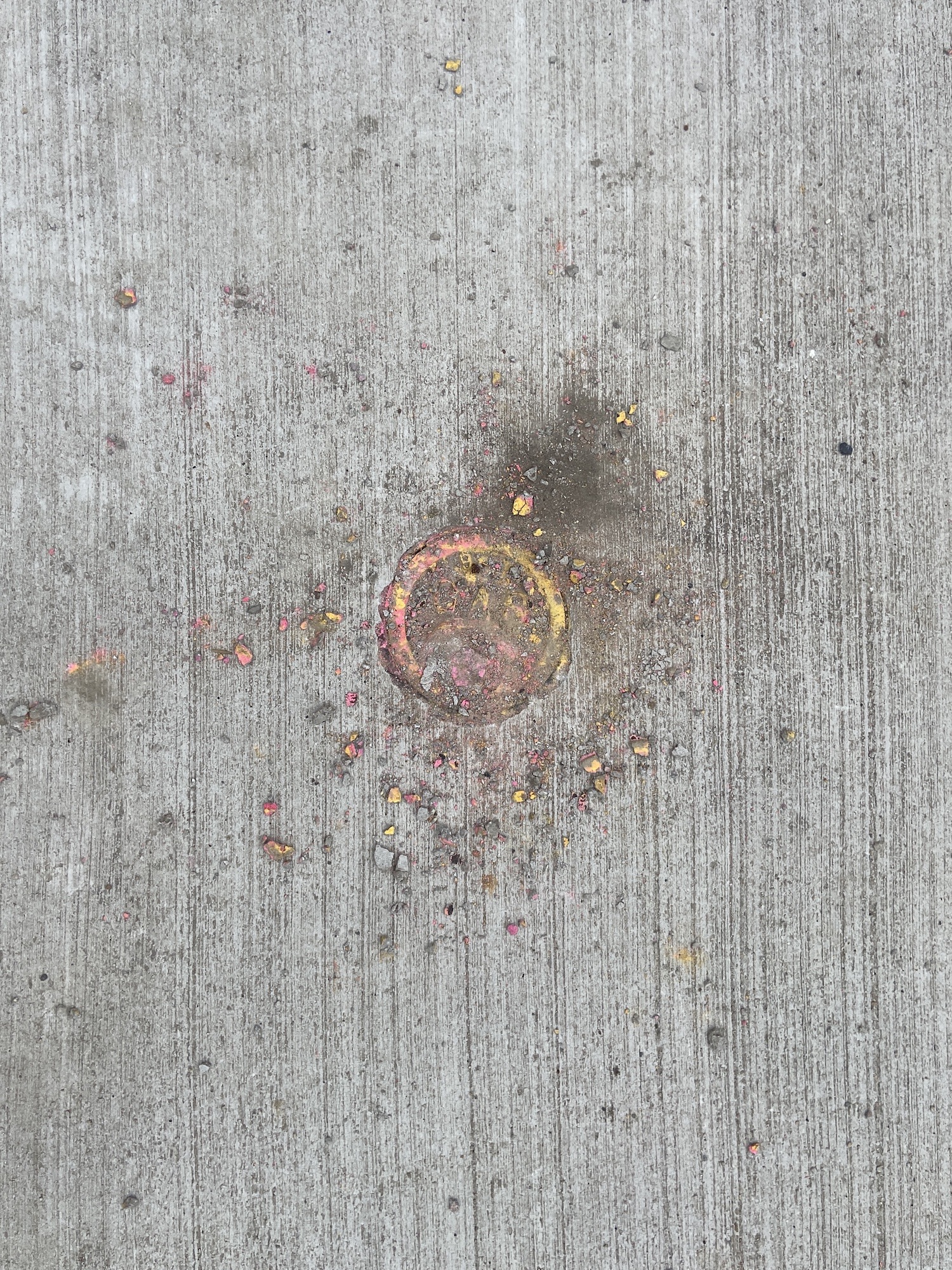 A gas shutoff cap in the sidewalk with yellow and pink pastel debris around it.