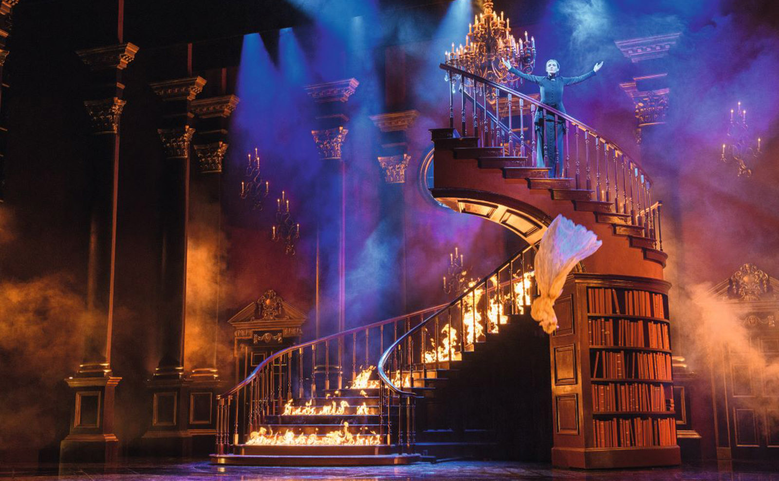 stage set for rebecca with a large staircase lined with bookshelves engulfed in flames