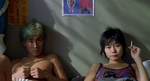 A screenshot from the movie 'Sympathy for Mr. Vengeance' of a young man with blue hair lying in bed with a young woman smoking a cigarette.