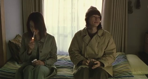 A screenshot of Teru sitting on a bed with a woman who is lighting a cigarette. From the movie 'Laundry'.