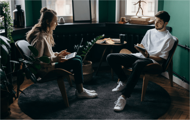 2 people sitting in a corner of a well-lit room, talking seriously to each other.