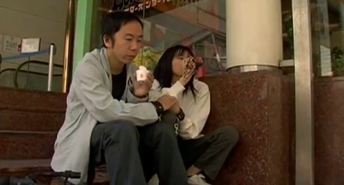 A screenshot from the horror film 'Marebito' of a man and young woman sitting on a set of steps outside of a shopping mall. The man is drinking an ice coffee and the girl is drinking a bottle of blood.