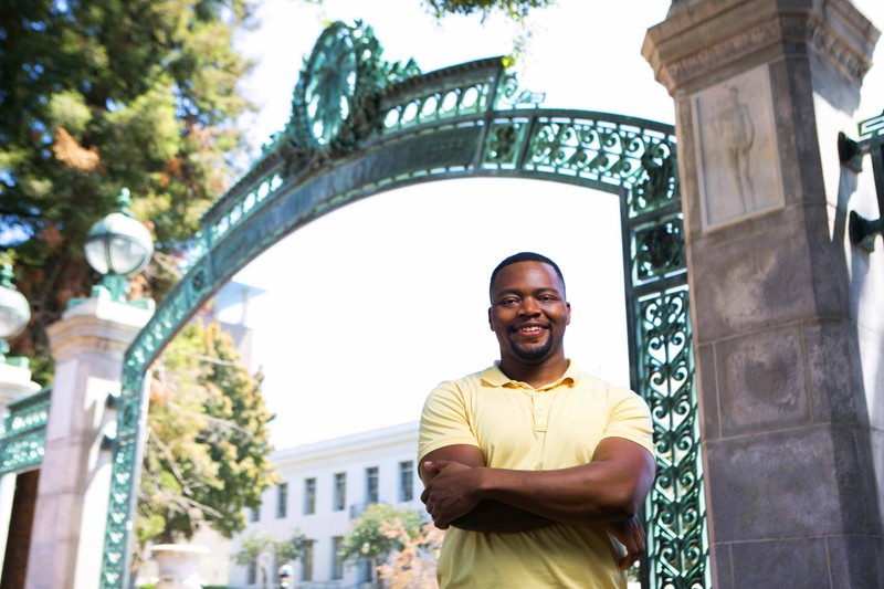 A smiling student in a yellow shirt stands in front of Sather Gate at the UC Berkeley