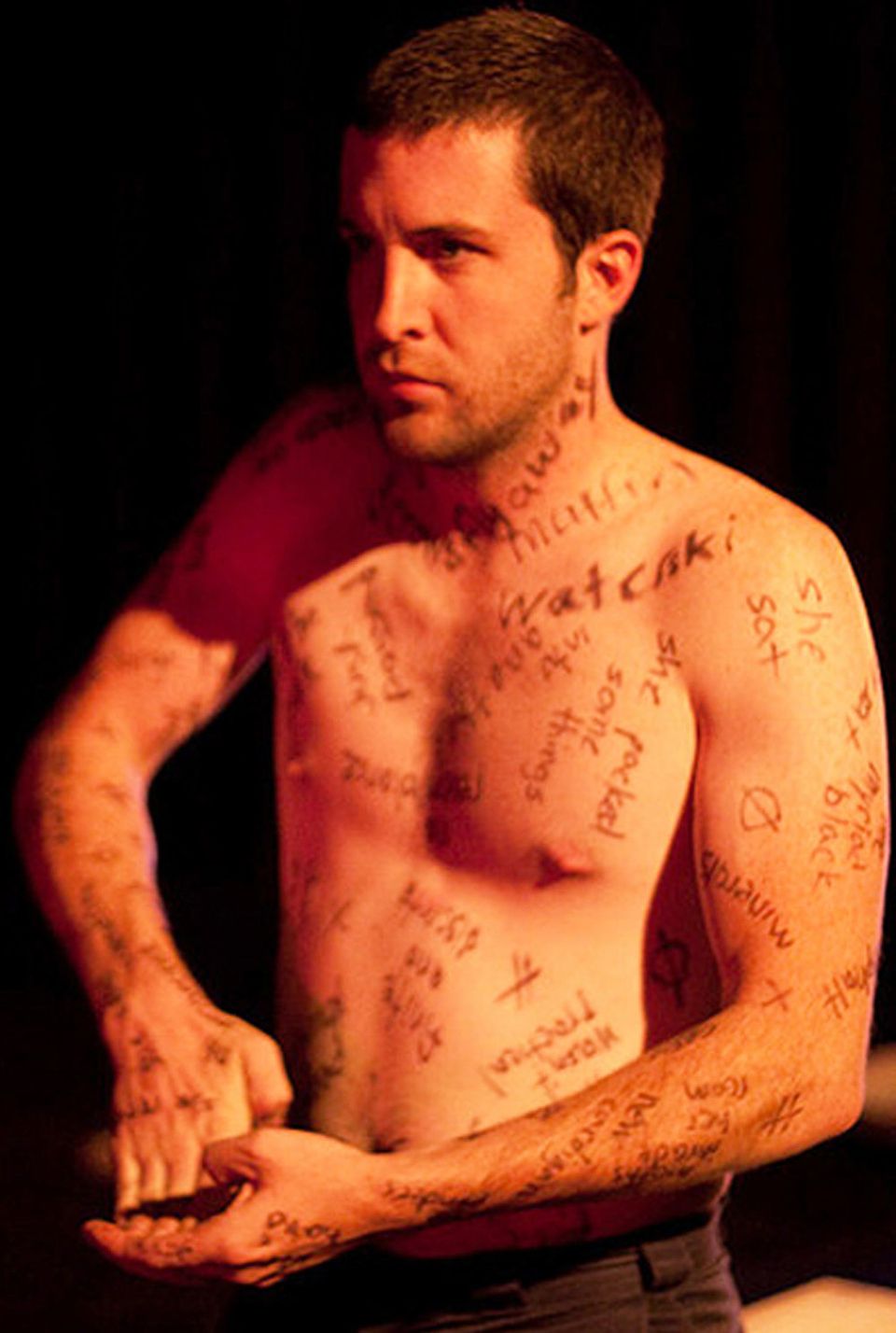 Shirtless man covered in writing, bends his fingers bakc