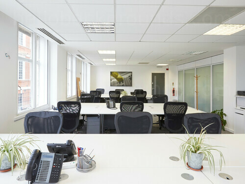 Simple shared office workspace with tiled ceiling above white desks with telephones and plants by black office chairs  Zoom background