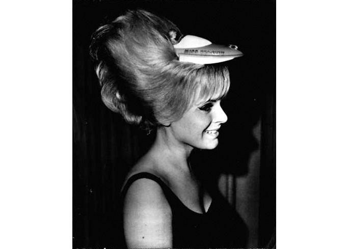  black and white photo of a blonde woman with a bouffant hairdo decorated with a toy airplane