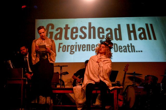 Jane reads a letter,
standing next to a disgusted Mrs Reed.
Behind them and the band, a projection reads
Gateshead Hall, forgiveness & death.
