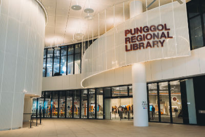 A photograph of the level 1 entrance of Punggol Regional Library. Clear floor-to-ceiling glass panels form the outer walls of the library, allowing the interior to be visible from the outside. The name of the library is displayed in large, bold brown letters on a white wall that protrudes from the second floor.