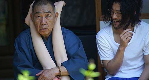 A screenshot from the movie 'Fumiko's Legs' of an old man with mannequin legs around his neck and a man sitting next to him laughing.