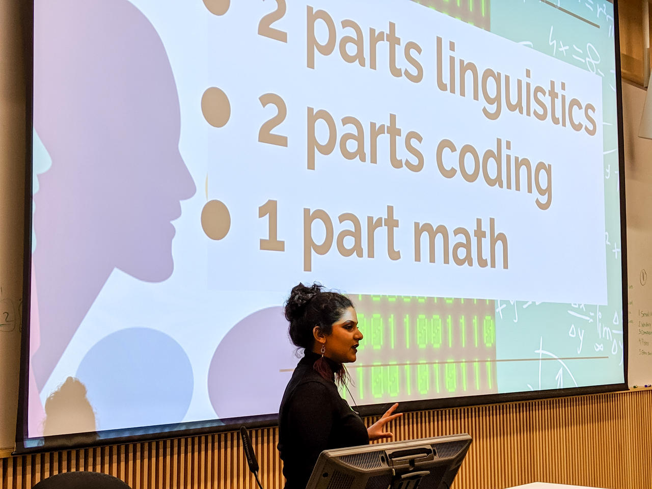 Vasundhara is standing and presenting in front of a screen where one of xyr slides is projected. The slide has three bullet points that say: 2 parts linguistics, 2 parts coding and 1 part math, which is xyr formula for xyr personal sweet spot with computational linguistics.