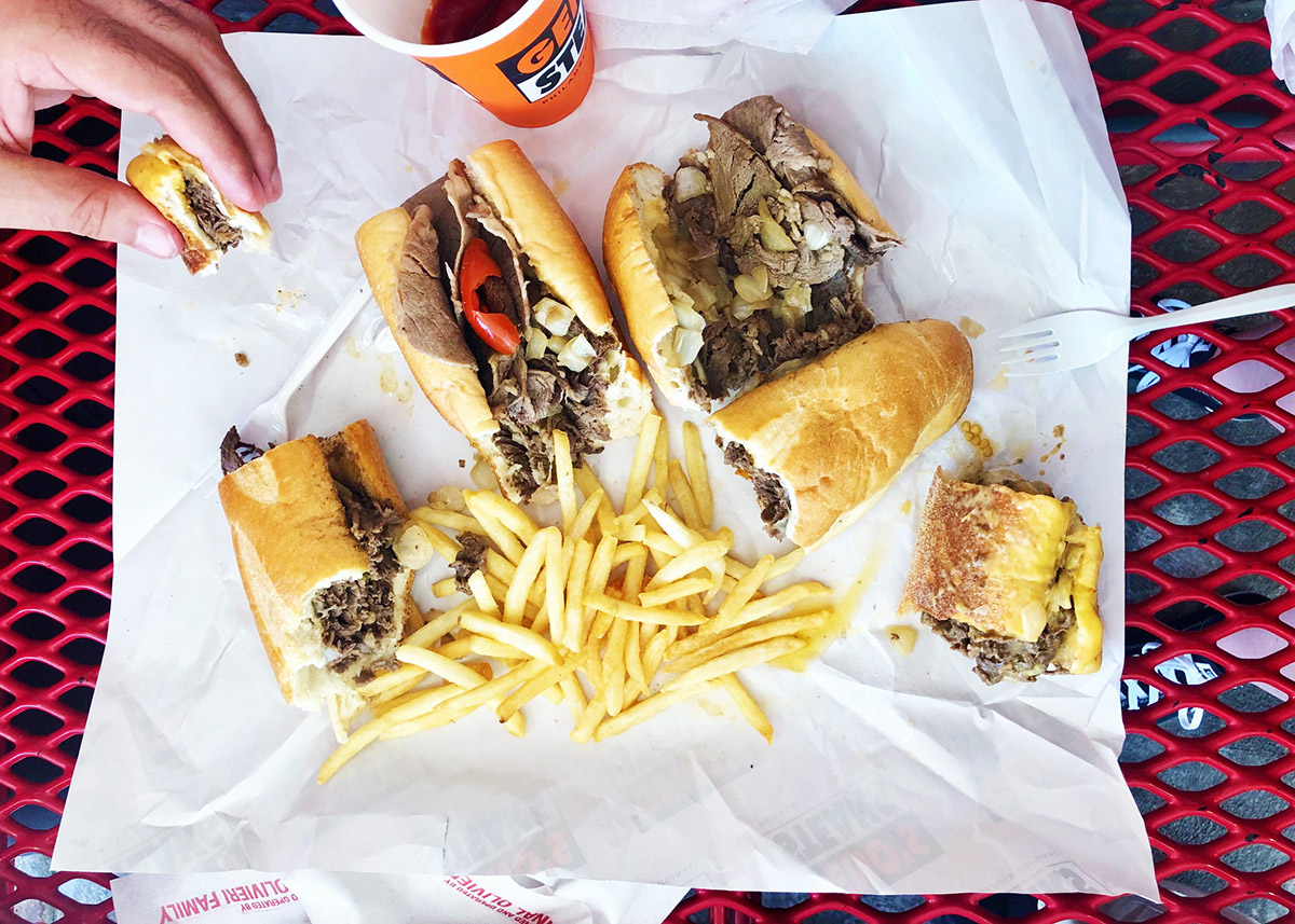 overhead view of multiple cheesesteak sandwiches on a red table with a pile of french fries