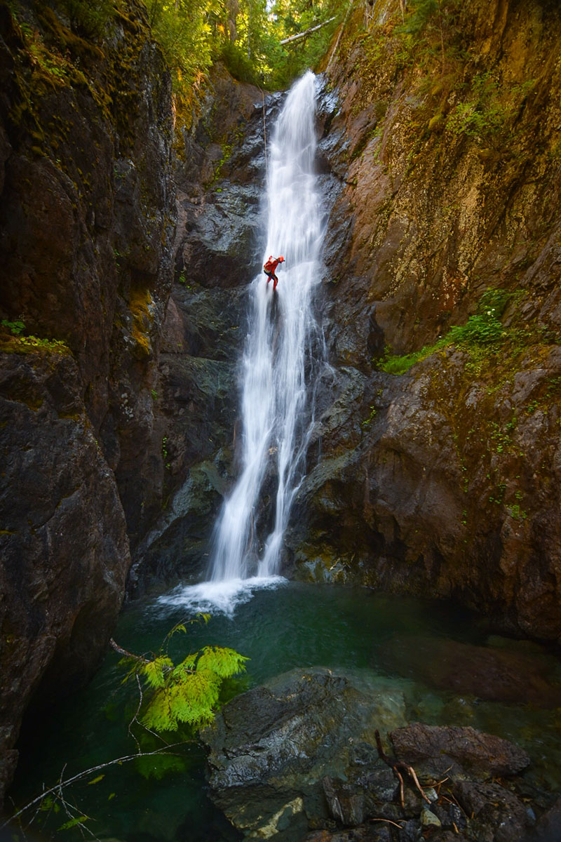 Man in red wetsuit climbing up a rockface in the middle of a long waterfall. The waterfall is in a deep canyon in a forest