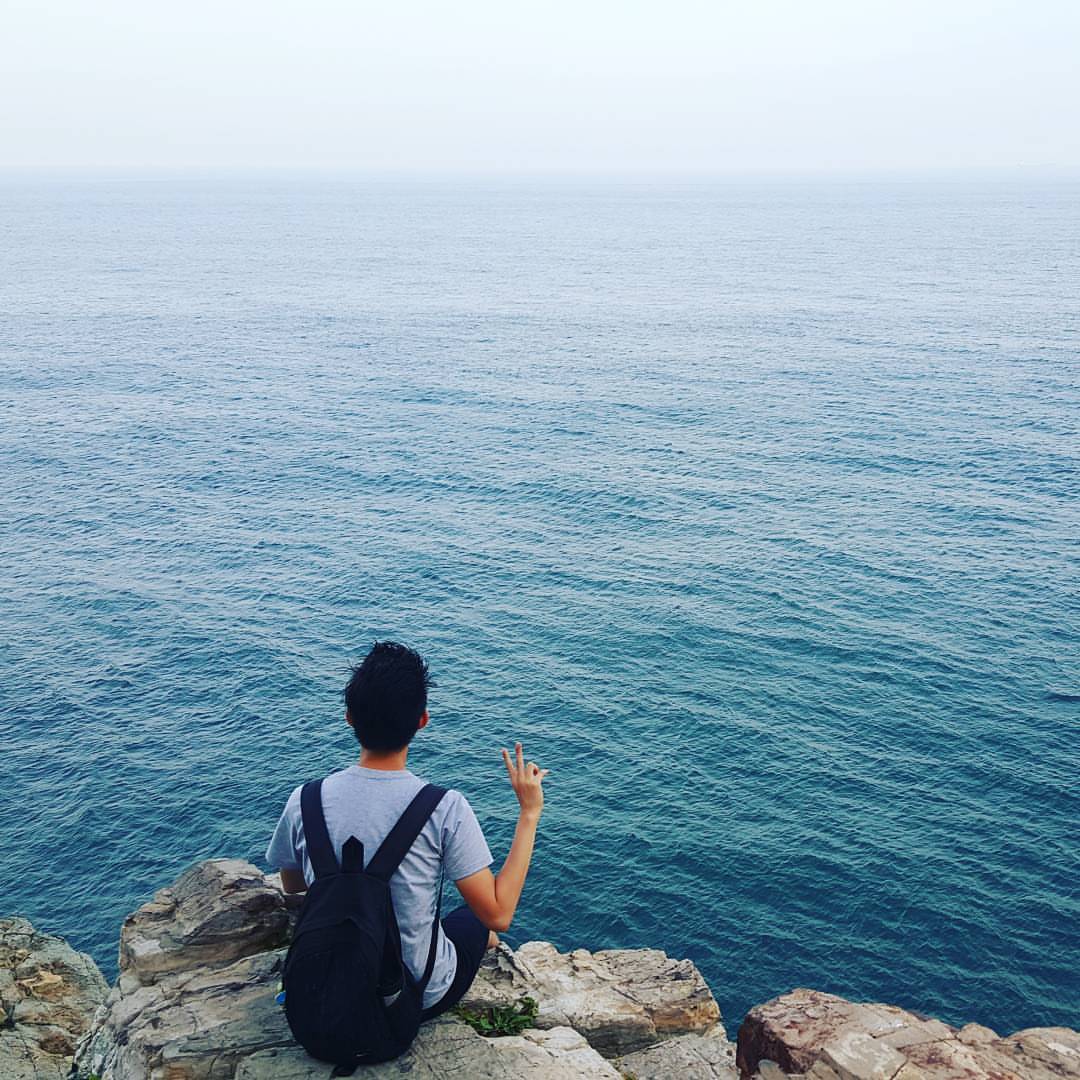 드넓은 바다를 바라보며 바위 절벽에 앉아 있는 엔디의 등 뒤에서 찍은 사진. 그의 오른손으로 V 포즈를 하고 있습니다.