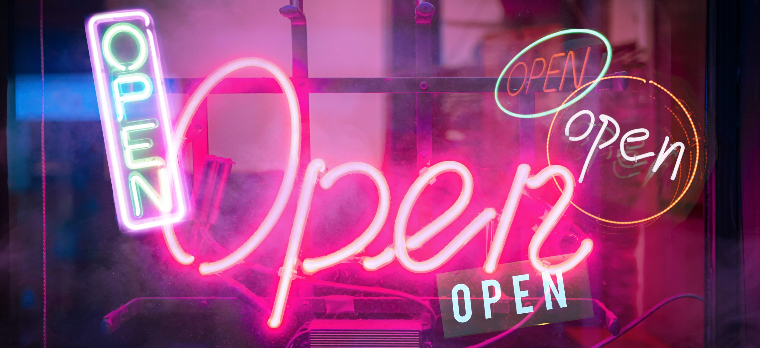 A photo illustration of several neon &quot;Open&quot; signs alight in the dark to signify re-opening of society after COVID-19