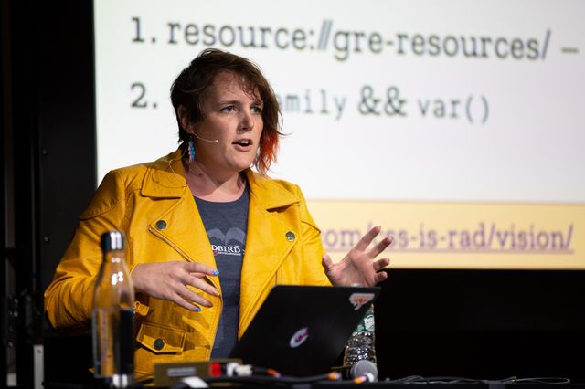 Miriam with a laptop and water bottle,
wearing a bright yellow leather jacket,
trans lightning earrings,
and a headset mic
while speaking at a conference.
