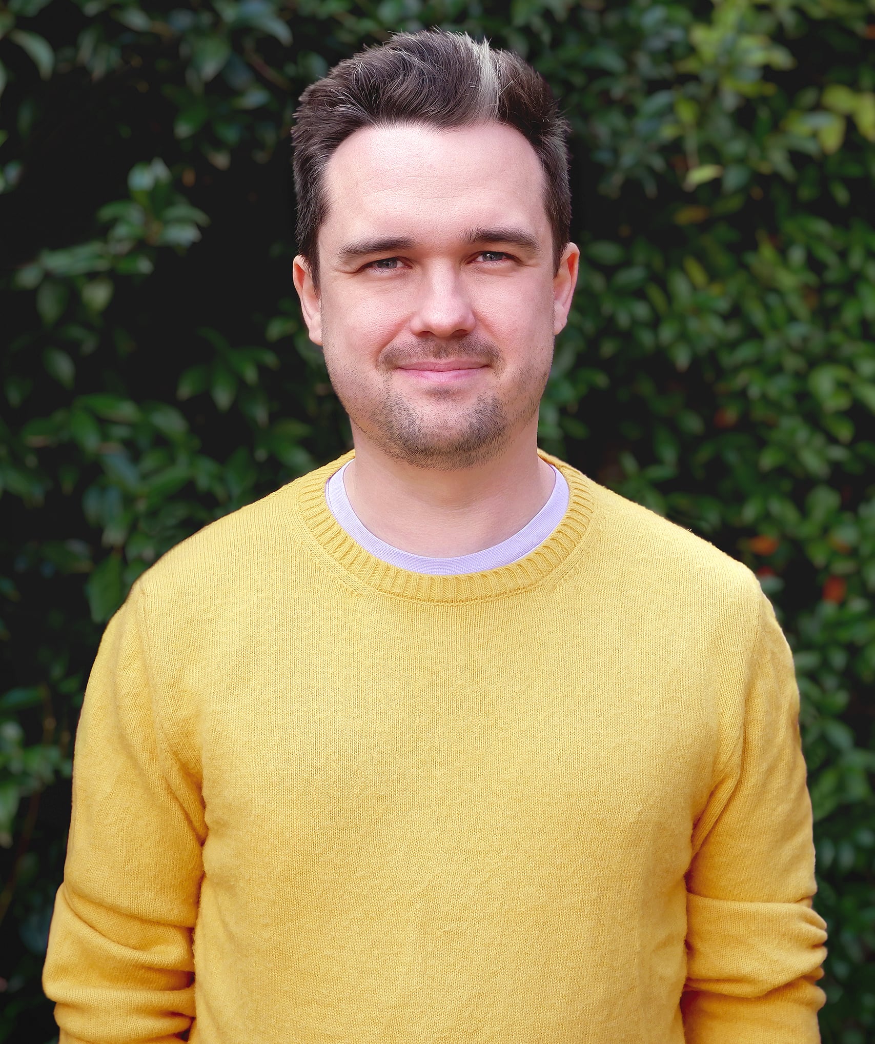 A picture of Elliot Midson wearing a yellow jumper in front of a green hedge.