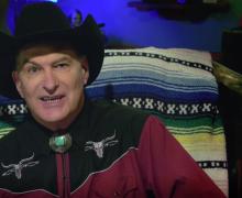 Joe Bob Briggs in Western attire with a bolo tie and Dwight Yoakam hat, arching his eyebrow at the camera.