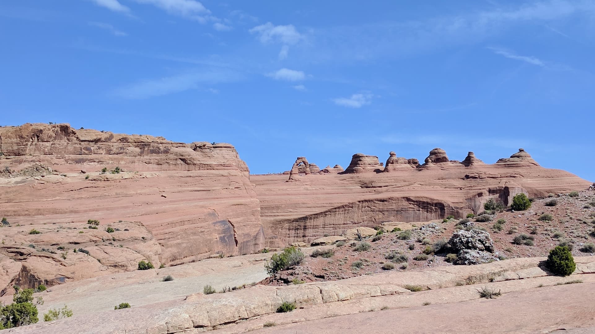 A distant view of Delicate Arch, which sits atop a large, wide sandstone fin. Scores of hikers, almost too small to see at this distance, cluster around it.