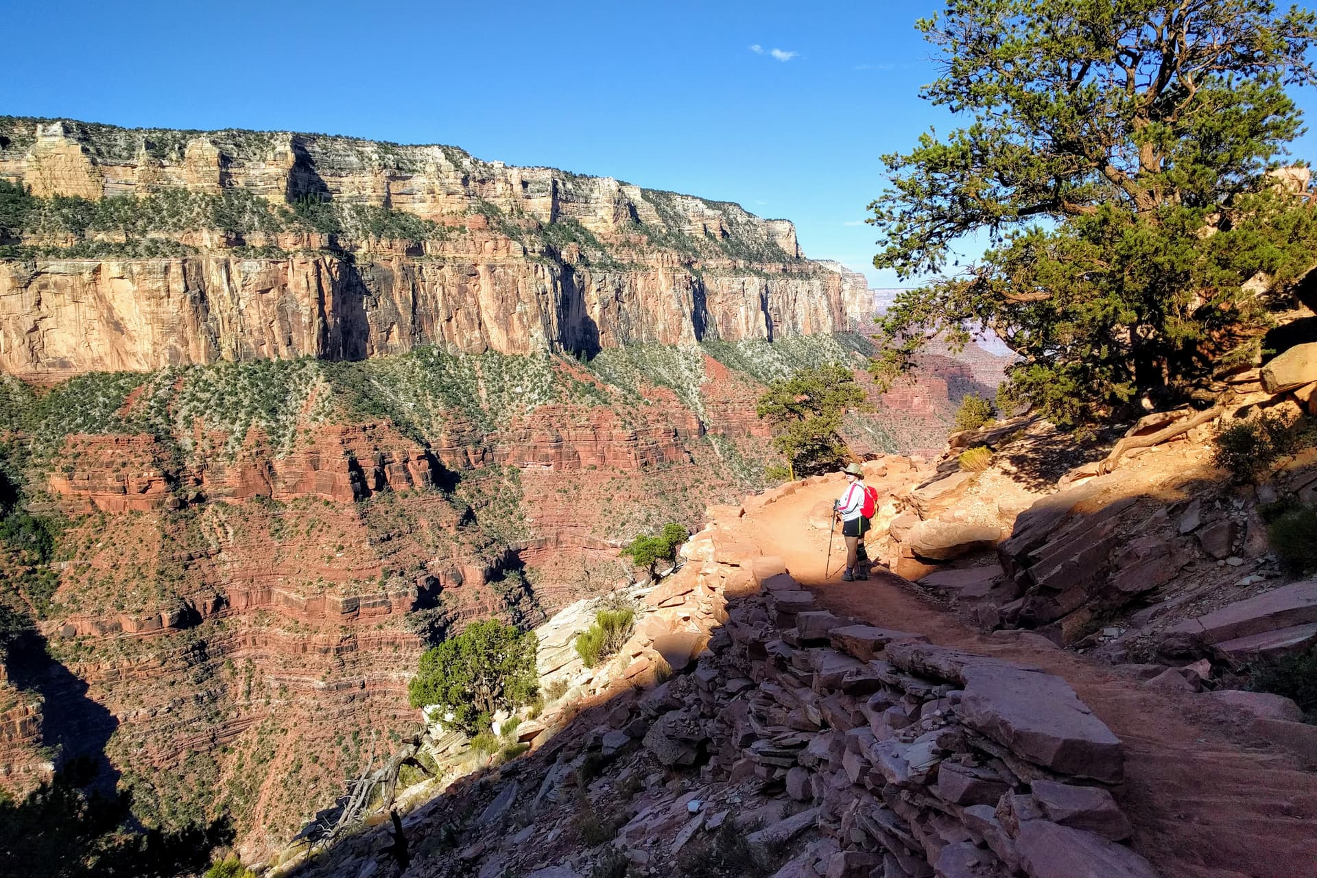 Len stands on a trail descending into the Grand Canyon from the South Rim. The shadow of the South Rim ends just before her, and in the background another part of the South Rim, composed of thick red and white sandstone bands, can be seen.