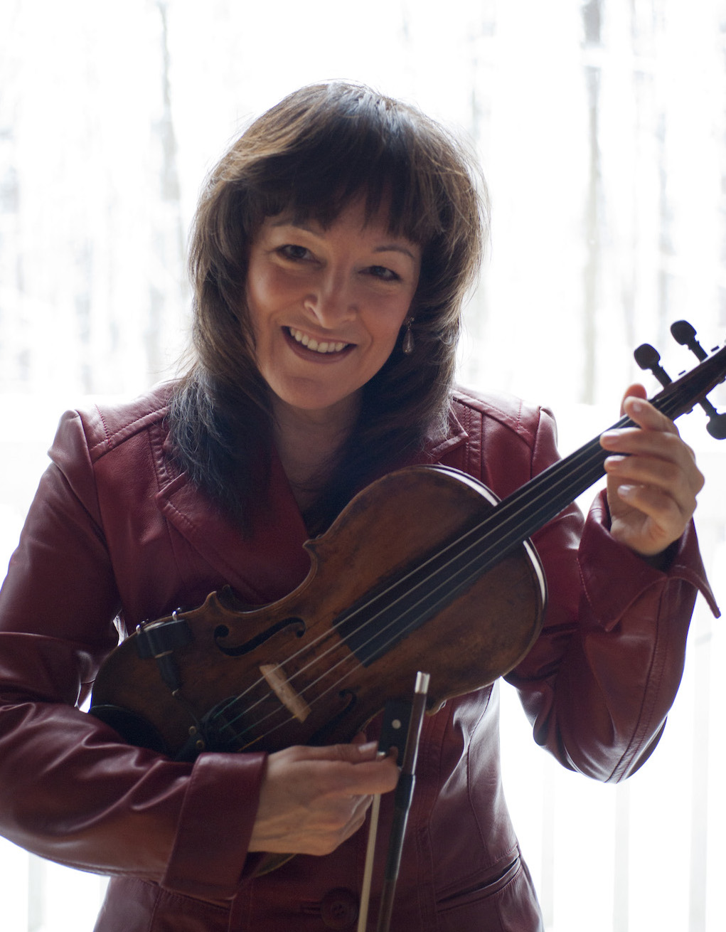 Cindy Thompson smiling and holding her fiddle and bow wearing a fancy red leather jacket.