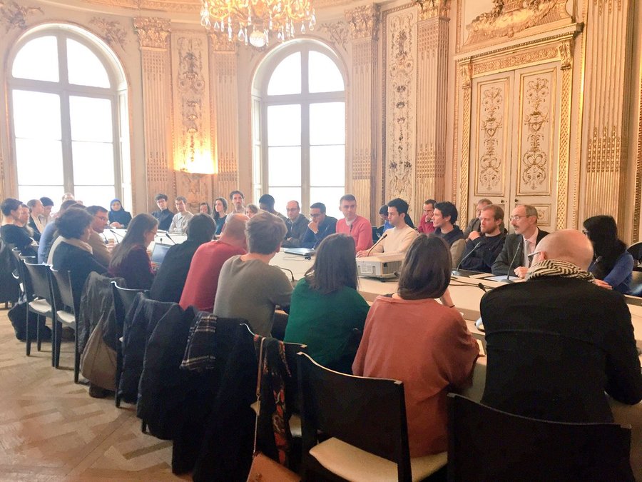 Plan d'ensemble d'une quarantaine de personnes autour d'une table, dans une salle de réunion agrémentée de dorures.