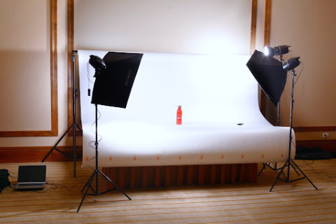 A small, table-top product photography studio with white backdrop and three lights is set up in an empty hotel conference room.
