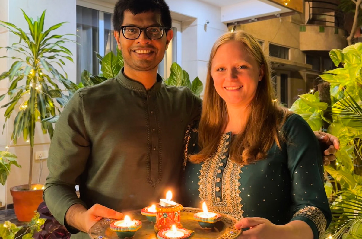 My fiance and I holding a tray of candles in front of house decorated with lights. We are wearing traditional Diwali shirts in tones of dark green.
