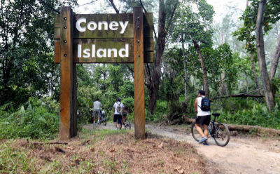A wooden sign board on Coney Island that reads 'Coney Island'. Cyclists walk past the sign with their bikes on a dirt path.