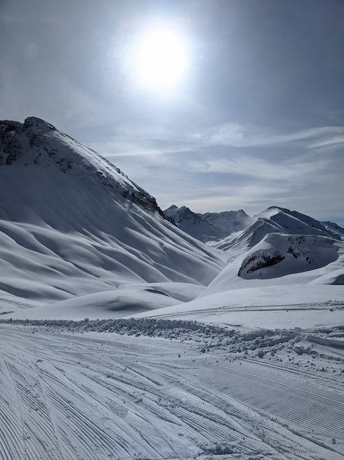 Groomed ski tracks in the foreground, sun behind mountains in the background