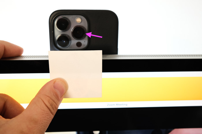 An Apple Studio Display with a MagSafe charger attached to the back holds an iPhone 13 and a person holds up a piece of paper to determine where the edge of the camera's field of view is