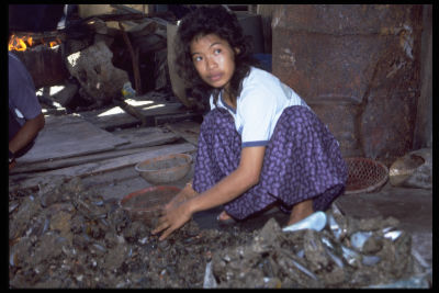 A female worker squatting down to deshell kupang (mussels) in a village house. A large cluster of kupang lies at her feet.