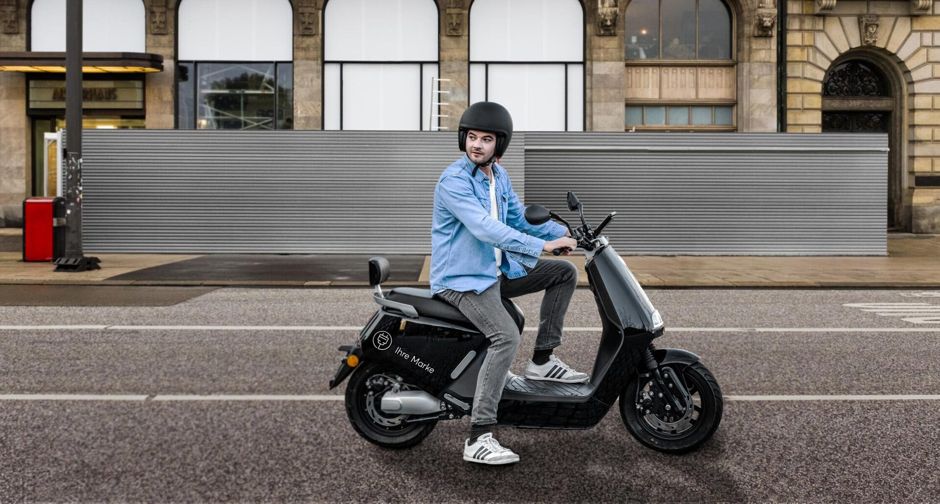 A caucasian man sitting on an e-moped in the middle of an urban street and looking sideways with one foot on the ground.