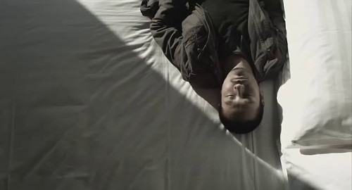 A screenshot from the film 'Here, Then', of a man lying on a bed, seen from above.