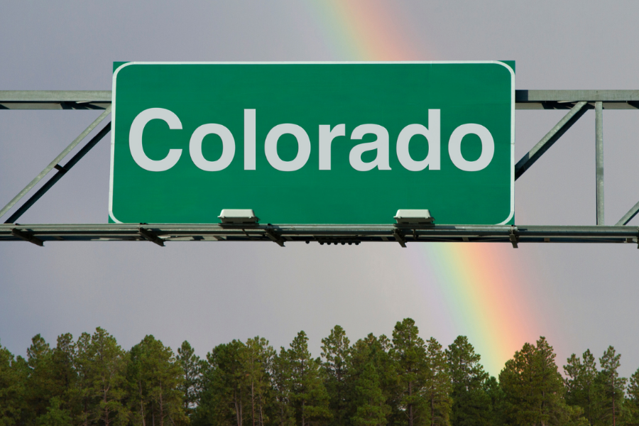 Billboard with white text reading 'Colorado' on green background. Behind the billboard are trees, and a rainbow in the sky.